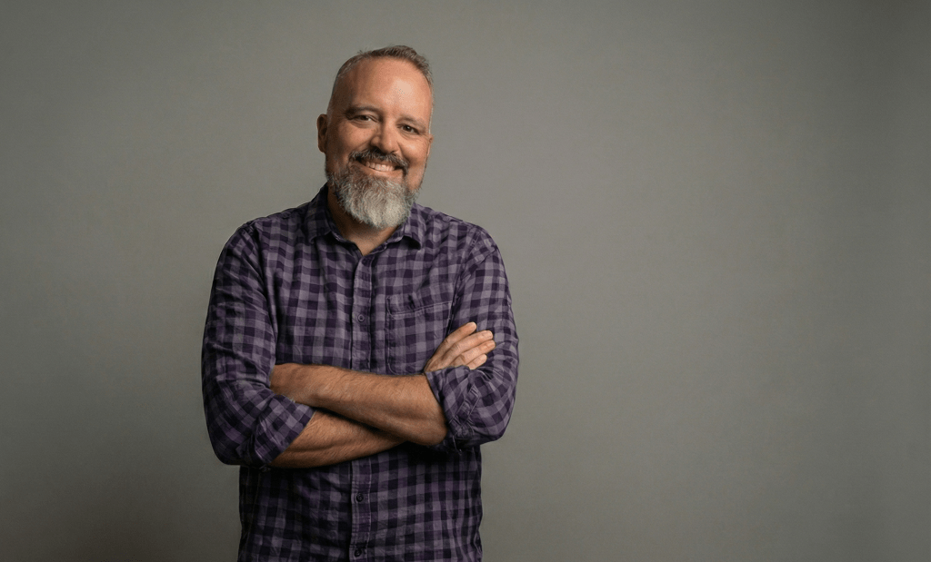 Coté with a gray beard and arms crossed wears a purple plaid shirt against a gray background, slightly offset.